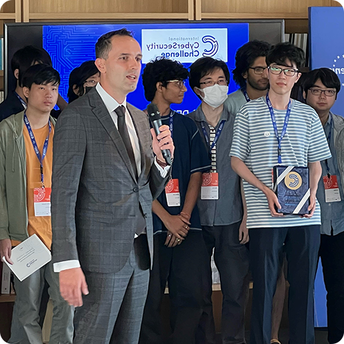 A host at the International Cybersecurity Challenge with a bunch of contestants behind him